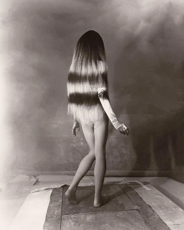 Monochrome photograph of a person with long, horizontally striped hair wearing gloves and minimal clothing posing in a studio environment.