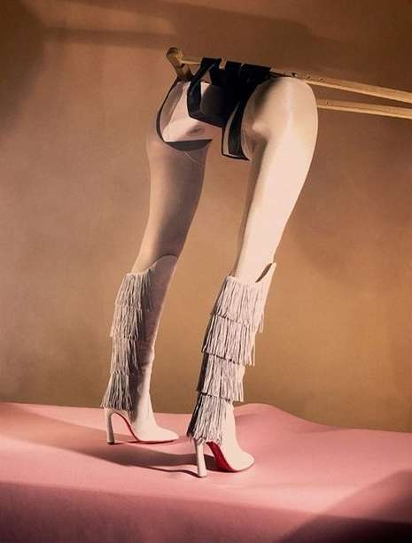 Artistic studio shot of a pair of white high-heeled boots with layered fringe, presented on a pink surface against a tan backdrop.