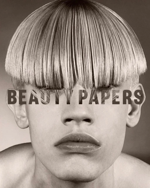 Close-up portrait of a person with a blunt blonde bowl cut hairstyle and the magazine title 'Beauty Papers' layered over the eyes.