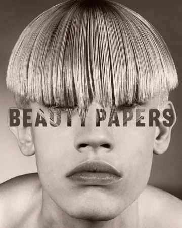 Close-up portrait of a person with a blunt blonde bowl cut hairstyle and the magazine title 'Beauty Papers' layered over the eyes.