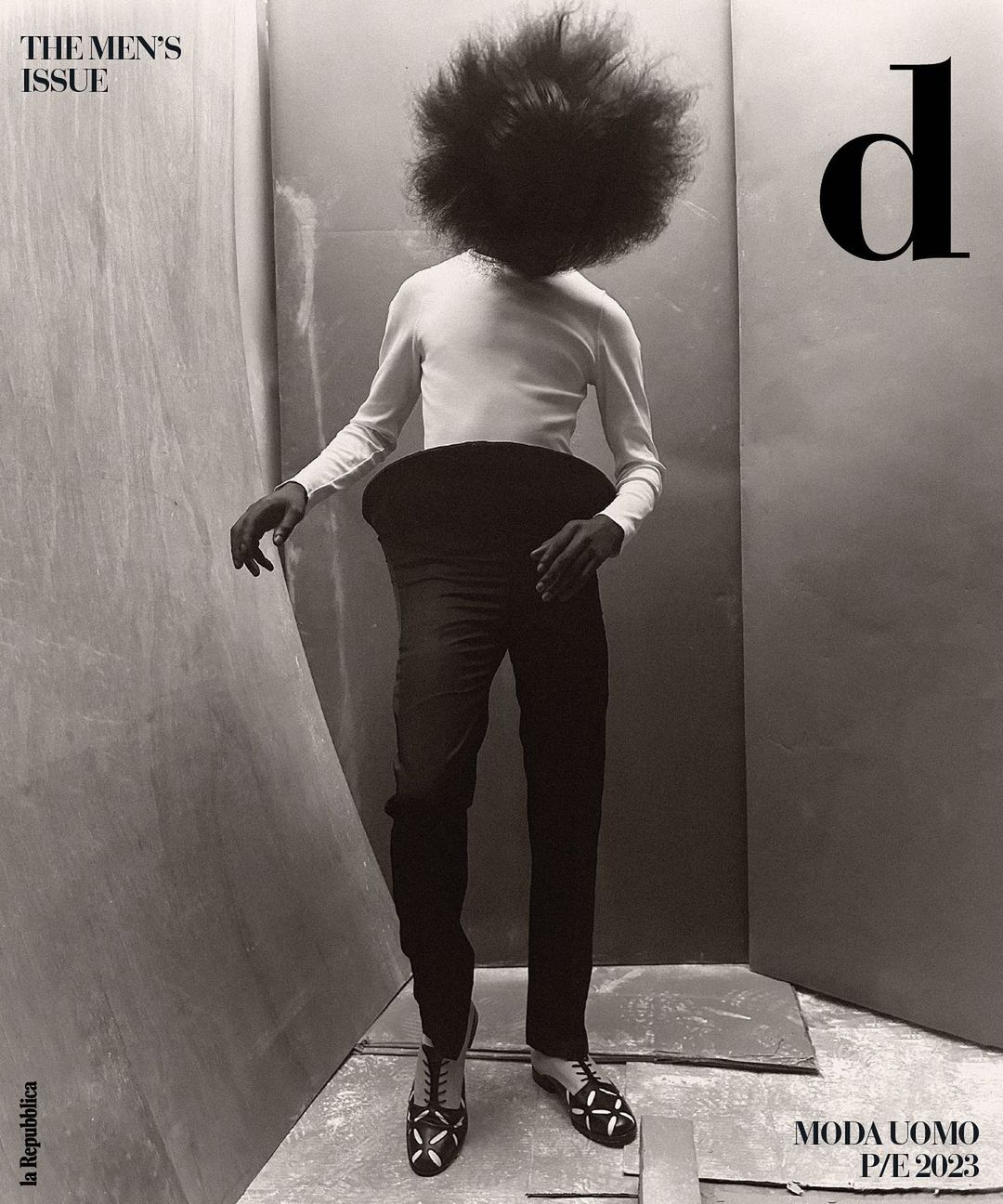 A person with large afro stands in black pants, white shirt, and patterned shoes against a simple studio backdrop for a men's fashion editorial.