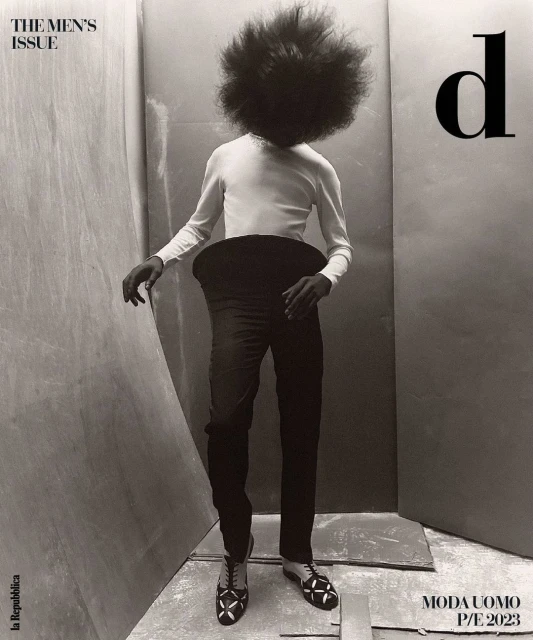 A person with large afro stands in black pants, white shirt, and patterned shoes against a simple studio backdrop for a men's fashion editorial.