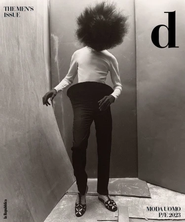 A person with large afro stands in black pants, white shirt, and patterned shoes against a simple studio backdrop for a men's fashion editorial.