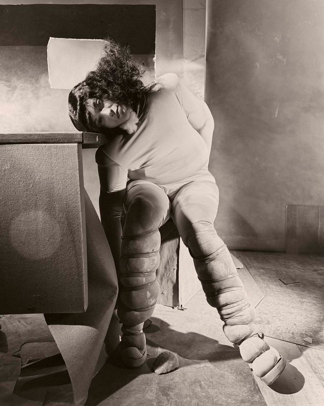 Artistic photograph of an adult female with unique leg padding, resting her head against a gray surface in a muted, indoor setting.