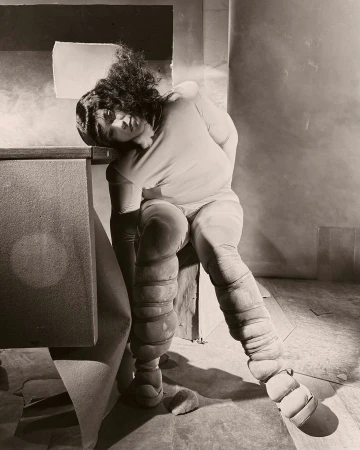Artistic photograph of an adult female with unique leg padding, resting her head against a gray surface in a muted, indoor setting.