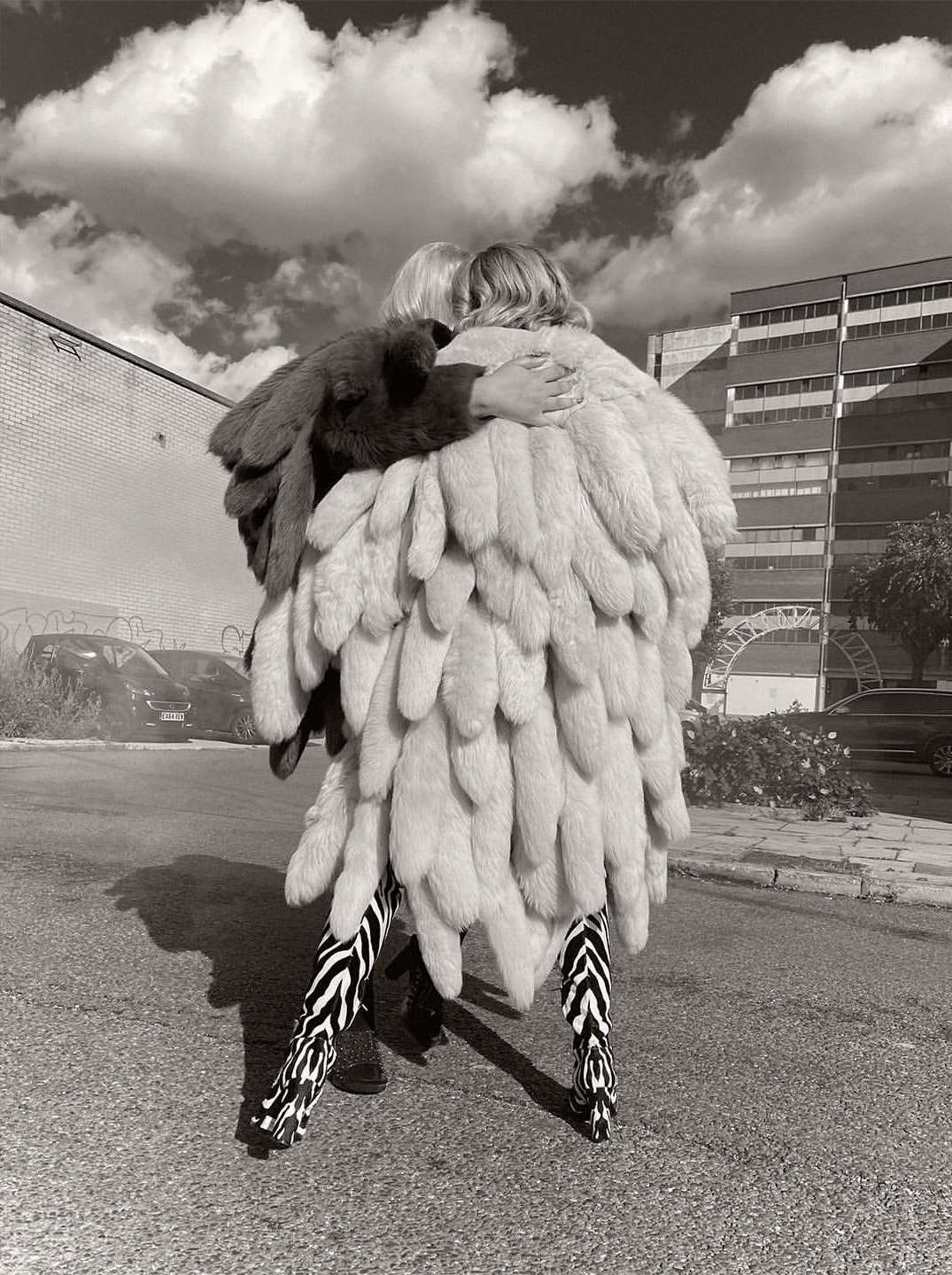 Two people in monochrome embrace, adorned in a striking feather coat and zebra-striped pants, against an urban backdrop and cloudy sky.