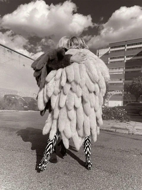 Two people in monochrome embrace, adorned in a striking feather coat and zebra-striped pants, against an urban backdrop and cloudy sky.