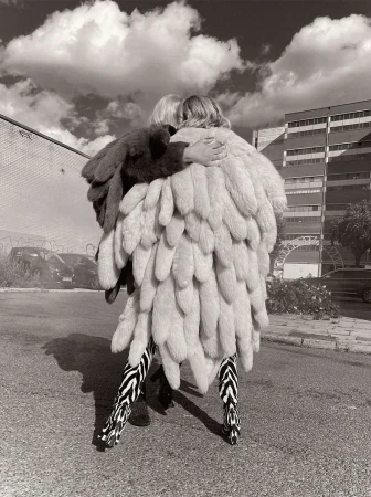 Two people in monochrome embrace, adorned in a striking feather coat and zebra-striped pants, against an urban backdrop and cloudy sky.