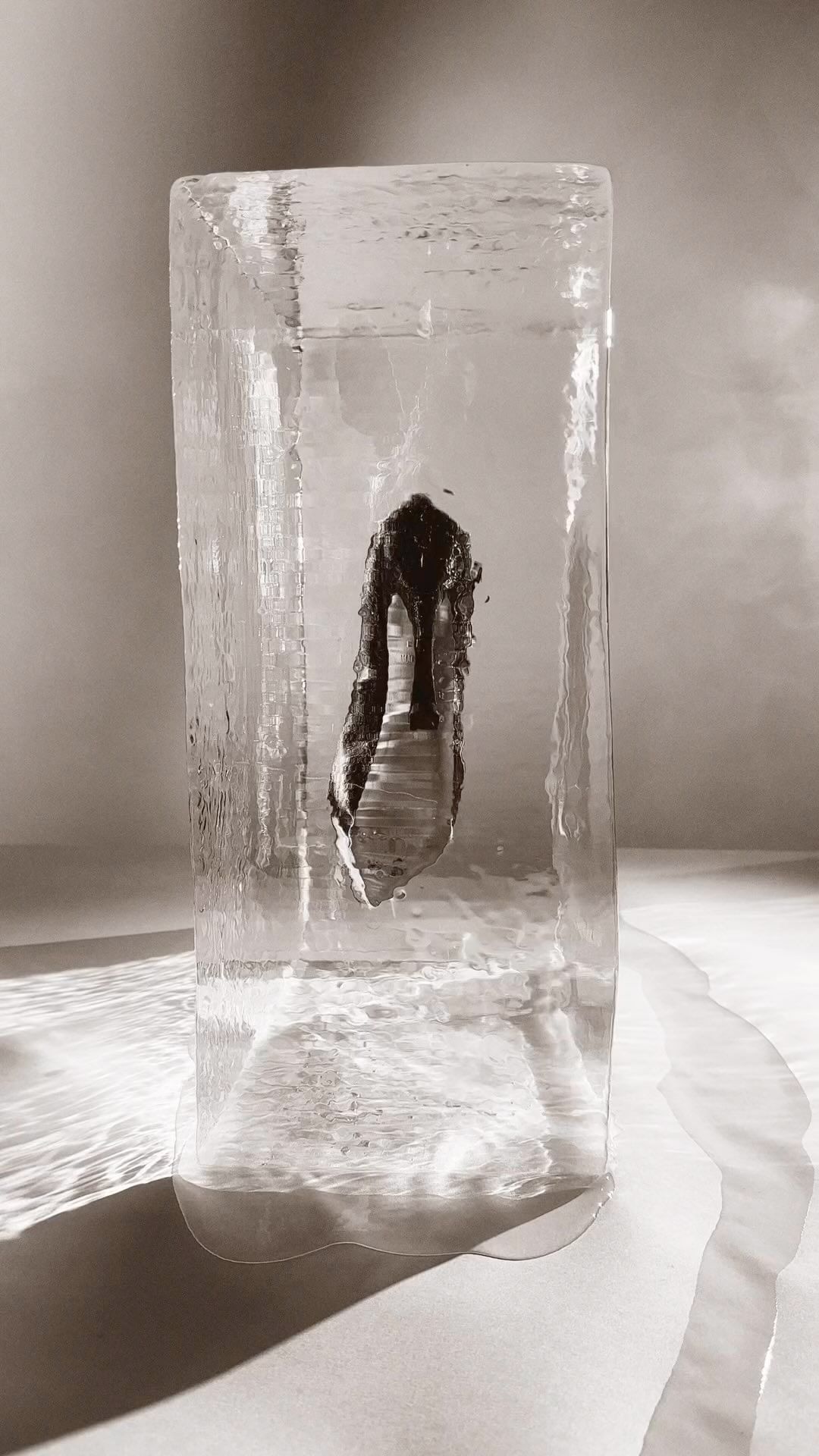 High heel pump encased in a rectangular clear ice block on a neutral surface with dramatic lighting creating shadows and a stark effect.