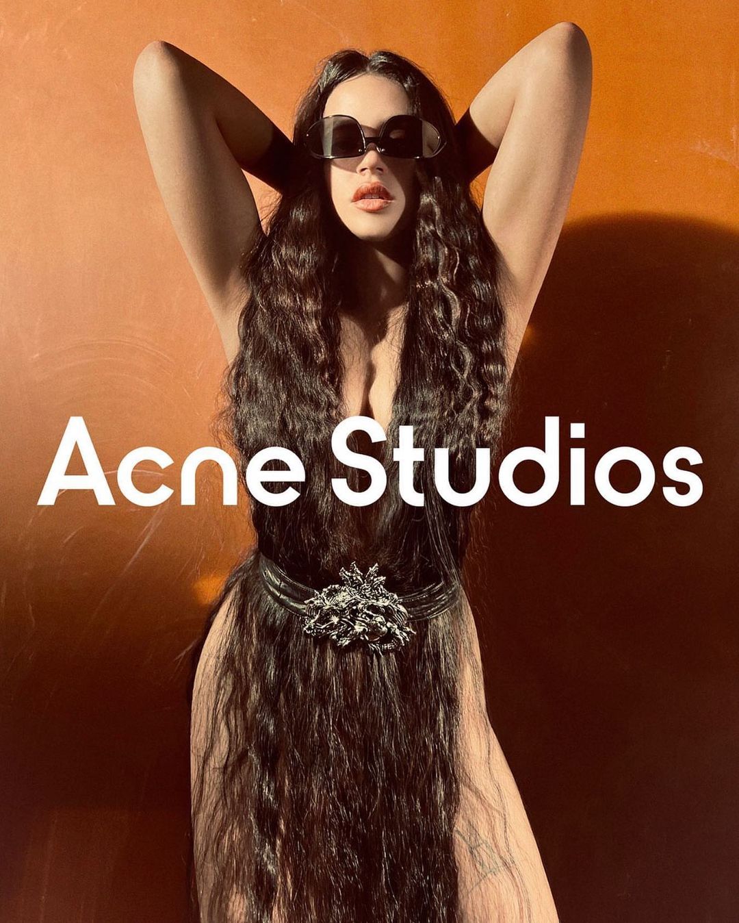 A woman with long dark hair stands with her hands behind her head in front of an orange background with the words Acne Studios overlaid.