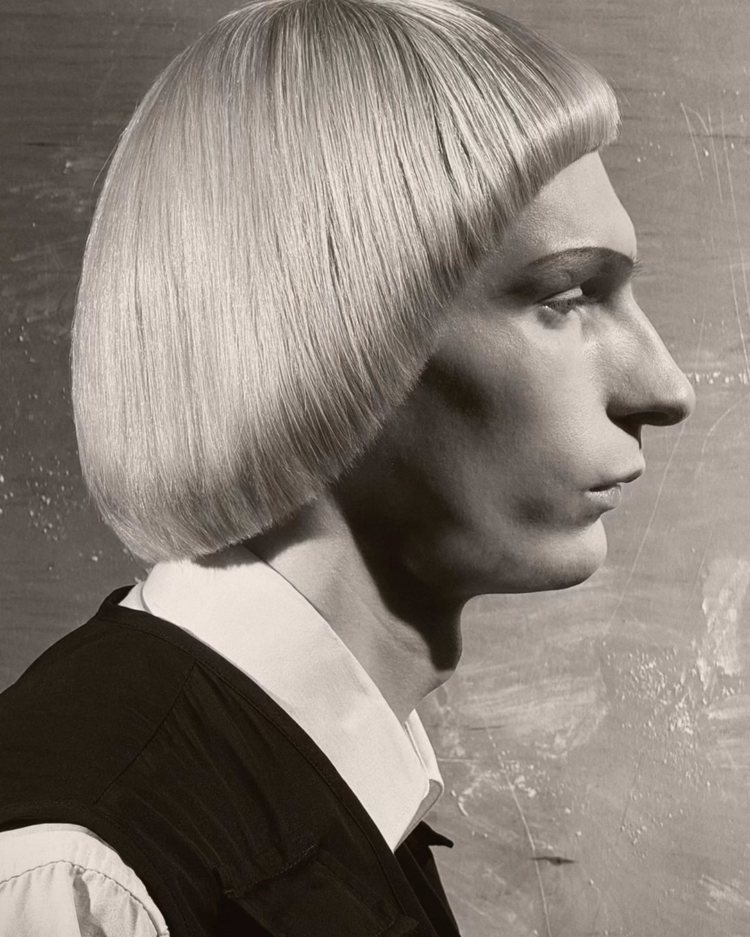 Monochrome portrait of a person in profile with a blunt blonde bowl cut, collared shirt, and black vest against a mottled background.