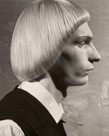 Monochrome portrait of a person in profile with a blunt blonde bowl cut, collared shirt, and black vest against a mottled background.