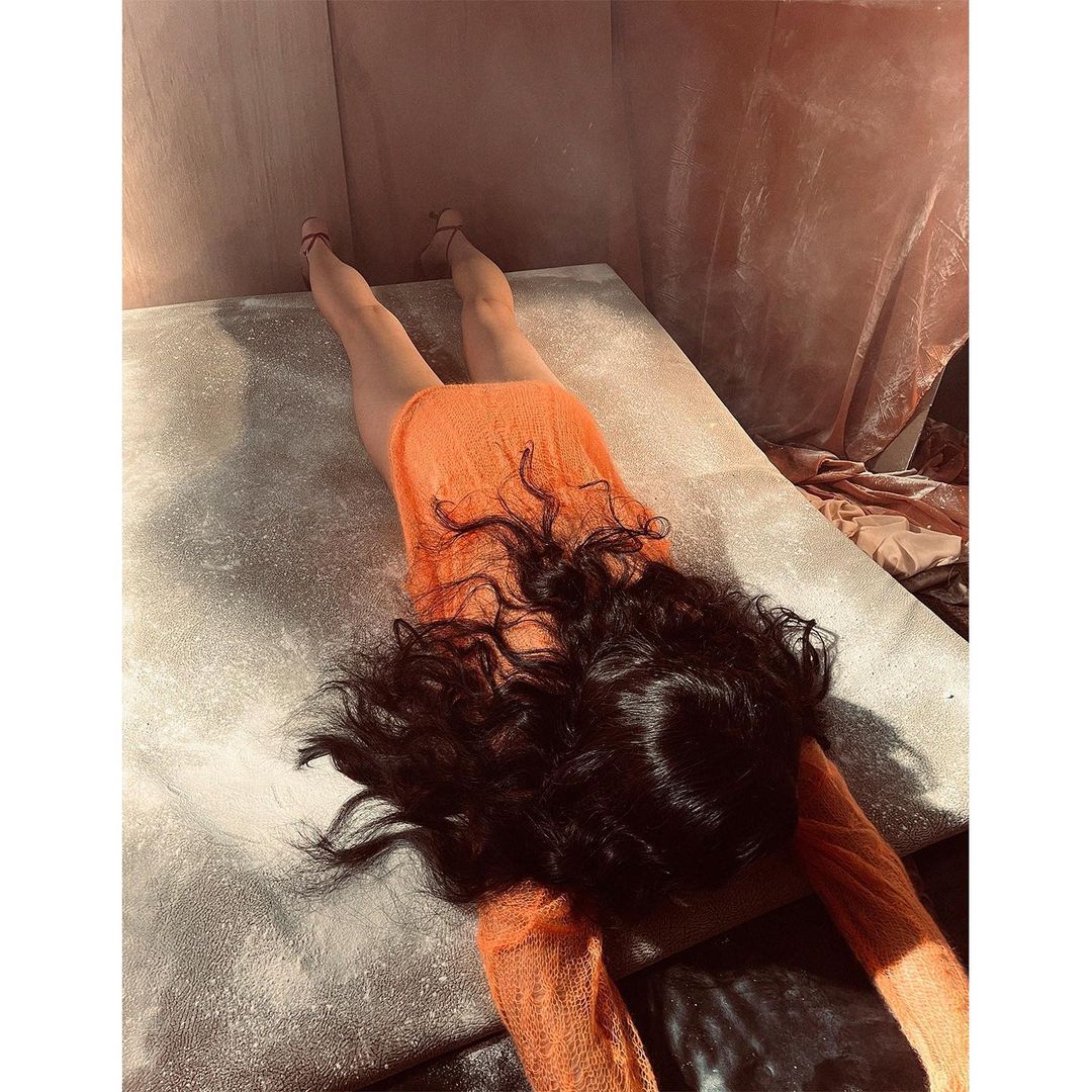 A prone adult woman with long dark hair in a bright-orange dress lies on a gray surface indoors, creating a striking, fashion-focused composition.