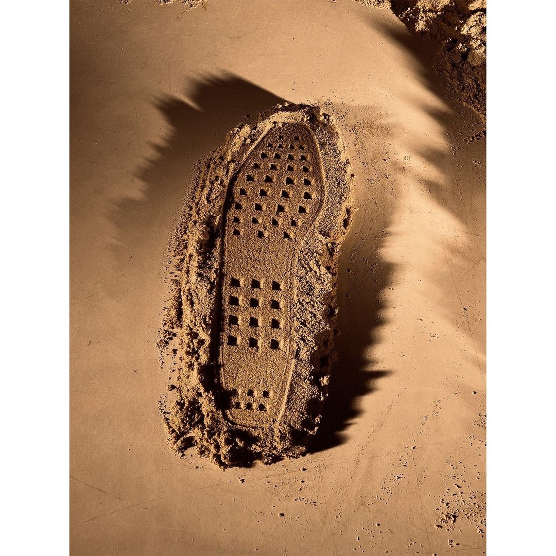 A close-up shows the impression of a shoe sole in sand, filled with a dark brown powder, set against a sandy surface.