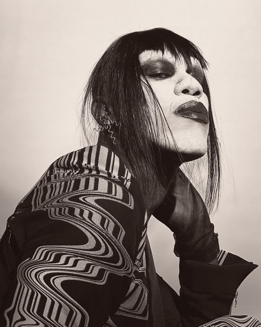 Striking portrait of a person with dark hair, theatrical makeup, and patterned jacket against a neutral backdrop.
