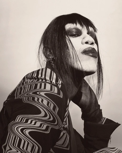 Striking portrait of a person with dark hair, theatrical makeup, and patterned jacket against a neutral backdrop.