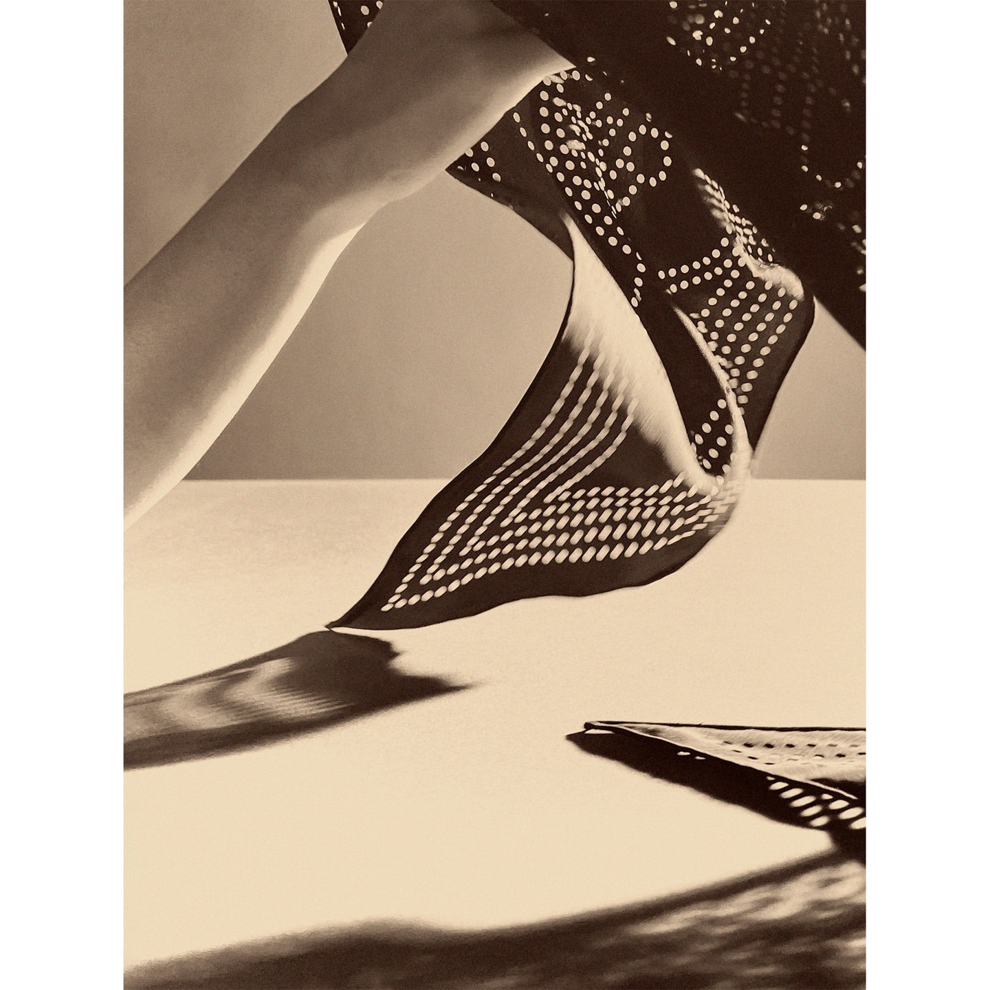 Monochrome photograph of a patterned scarf suspended in the air by an arm over a tabletop, showcasing shadows and textures.