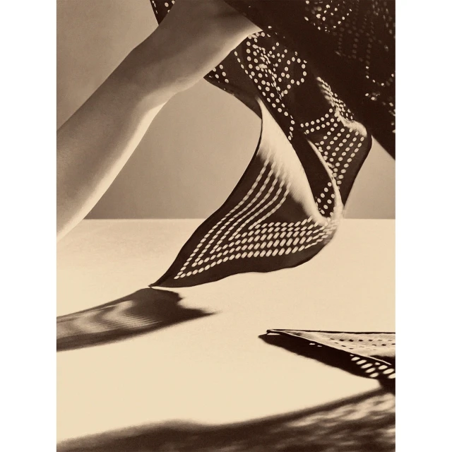 Monochrome photograph of a patterned scarf suspended in the air by an arm over a tabletop, showcasing shadows and textures.