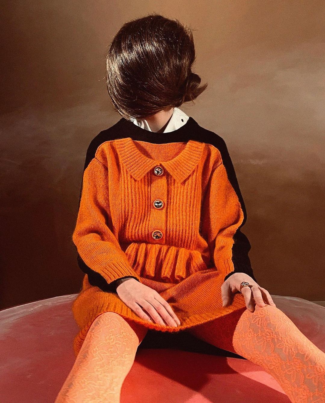A brunette model with orange dress and patterned tights poses in a studio with warm brown background, creating a retro fashion image.