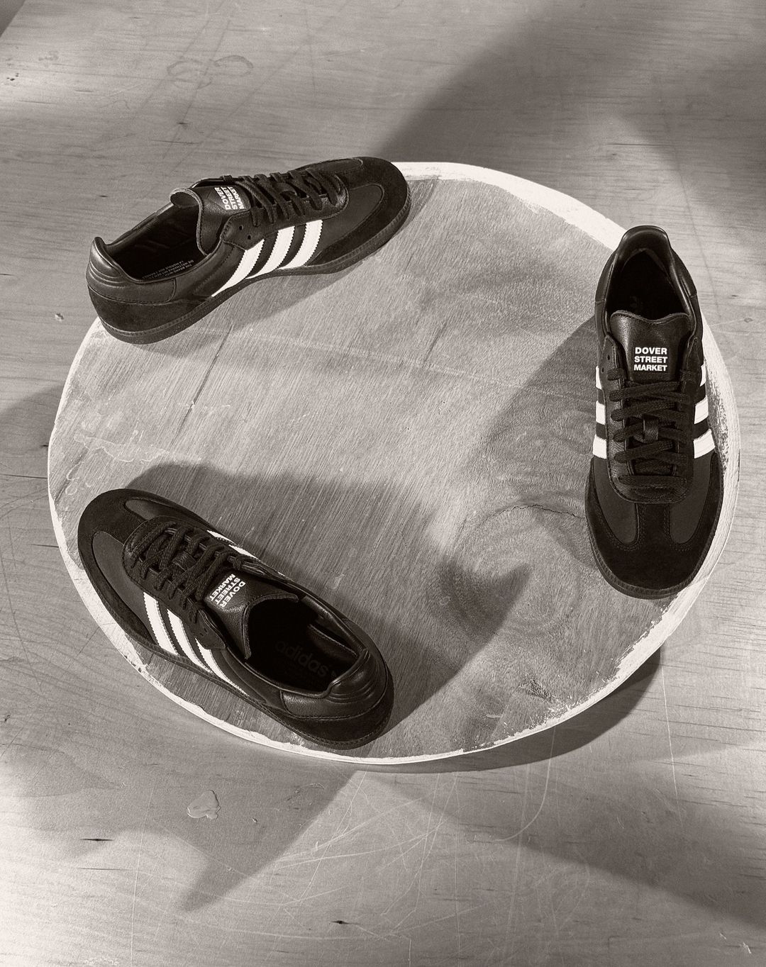 Three Adidas Samba sneakers arranged in a circular pattern on a light wooden surface, showcasing a minimalist shoe display.