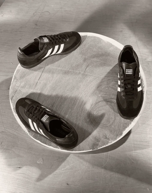 Three Adidas Samba sneakers arranged in a circular pattern on a light wooden surface, showcasing a minimalist shoe display.