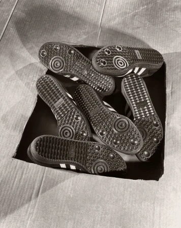 Multiple pairs of Adidas Samba sneakers in a cardboard box, soles facing up, featuring the brand's classic three-stripe design.