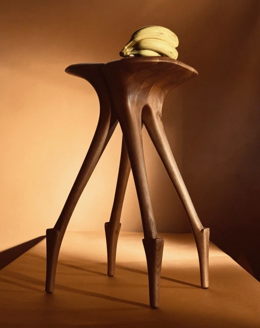 Elevated view of a modern wood stool with unique leg design, topped with a bunch of bananas against a warm, solid-colored backdrop.