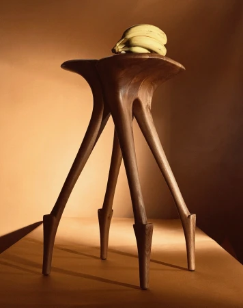 Elevated view of a modern wood stool with unique leg design, topped with a bunch of bananas against a warm, solid-colored backdrop.