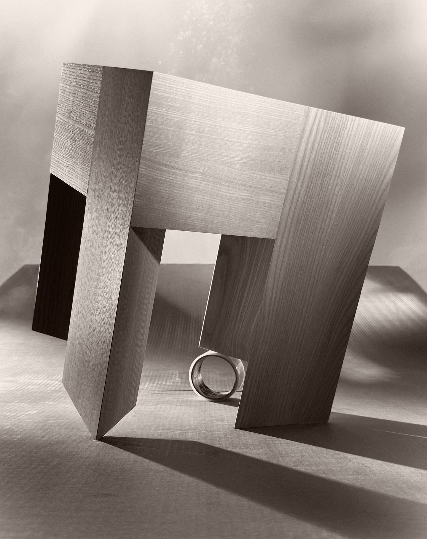 Abstract geometric wood sculpture with a metal ring, illuminated with studio lighting on a wooden surface, creating strong shadows.