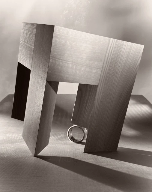 Abstract geometric wood sculpture with a metal ring, illuminated with studio lighting on a wooden surface, creating strong shadows.