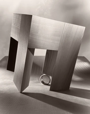 Abstract geometric wood sculpture with a metal ring, illuminated with studio lighting on a wooden surface, creating strong shadows.