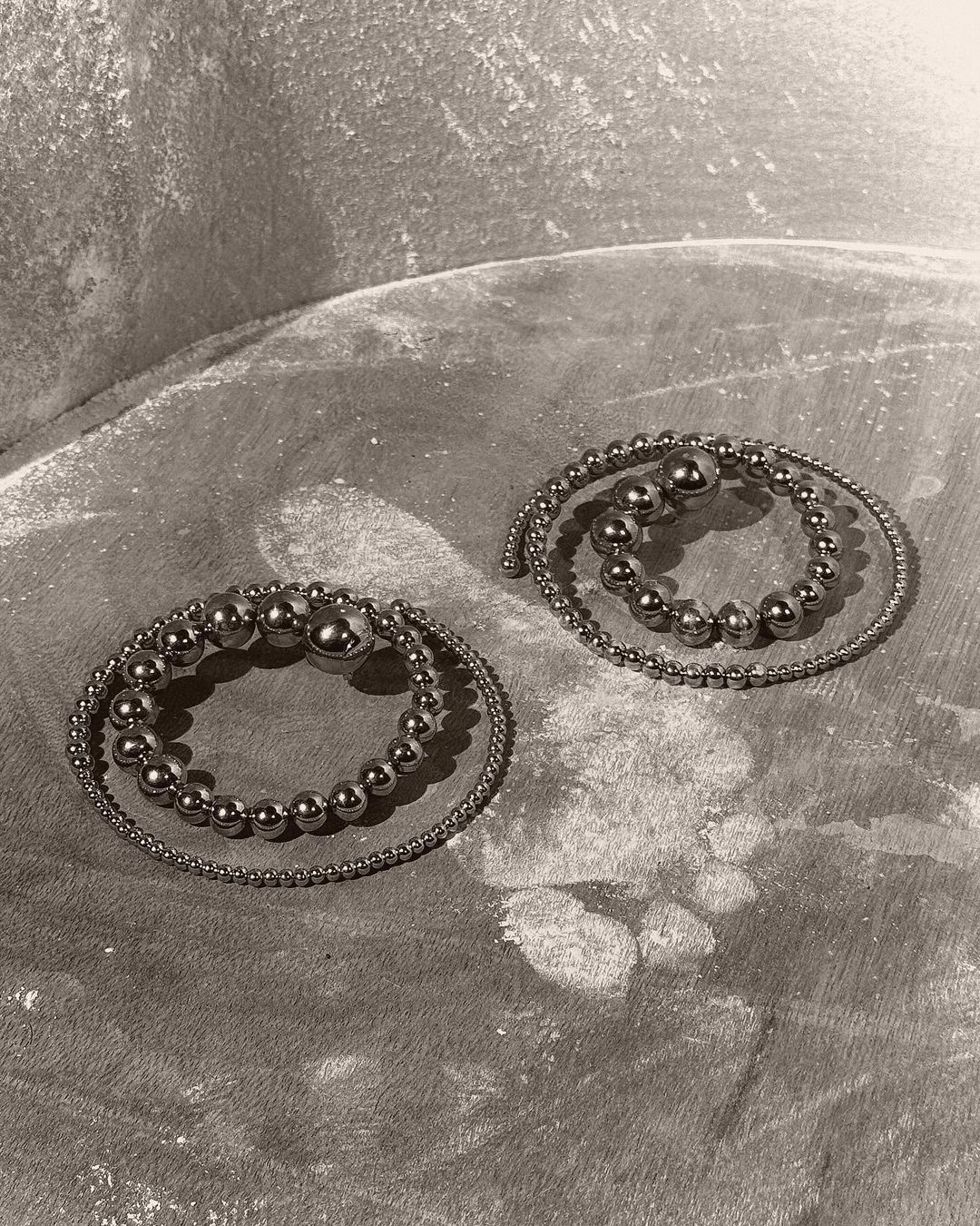 Two silver bead bracelets are coiled on a textured gray surface, creating a simple yet elegant composition in a studio setting.