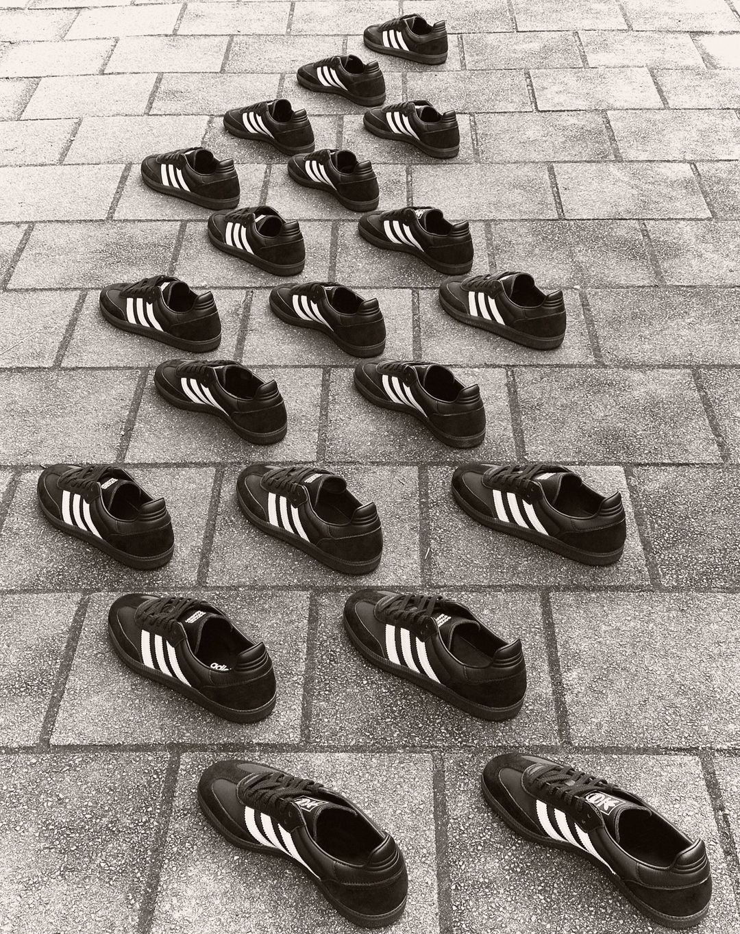 Multiple Adidas sneakers arranged in a pyramid pattern on a brick surface, creating a rhythmic and symmetrical composition, monochrome.