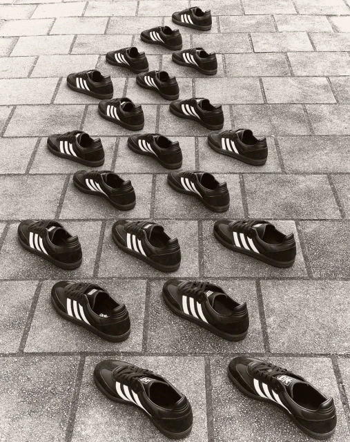 Multiple Adidas sneakers arranged in a pyramid pattern on a brick surface, creating a rhythmic and symmetrical composition, monochrome.