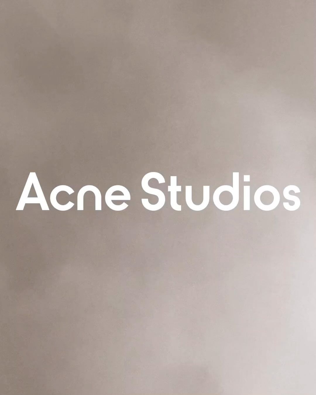 Minimalist studio shot of 'Acne Studios' brand name in bold white sans-serif typeface on a muted beige background with soft gradations.