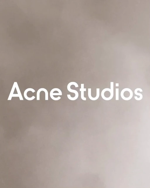 Minimalist studio shot of 'Acne Studios' brand name in bold white sans-serif typeface on a muted beige background with soft gradations.