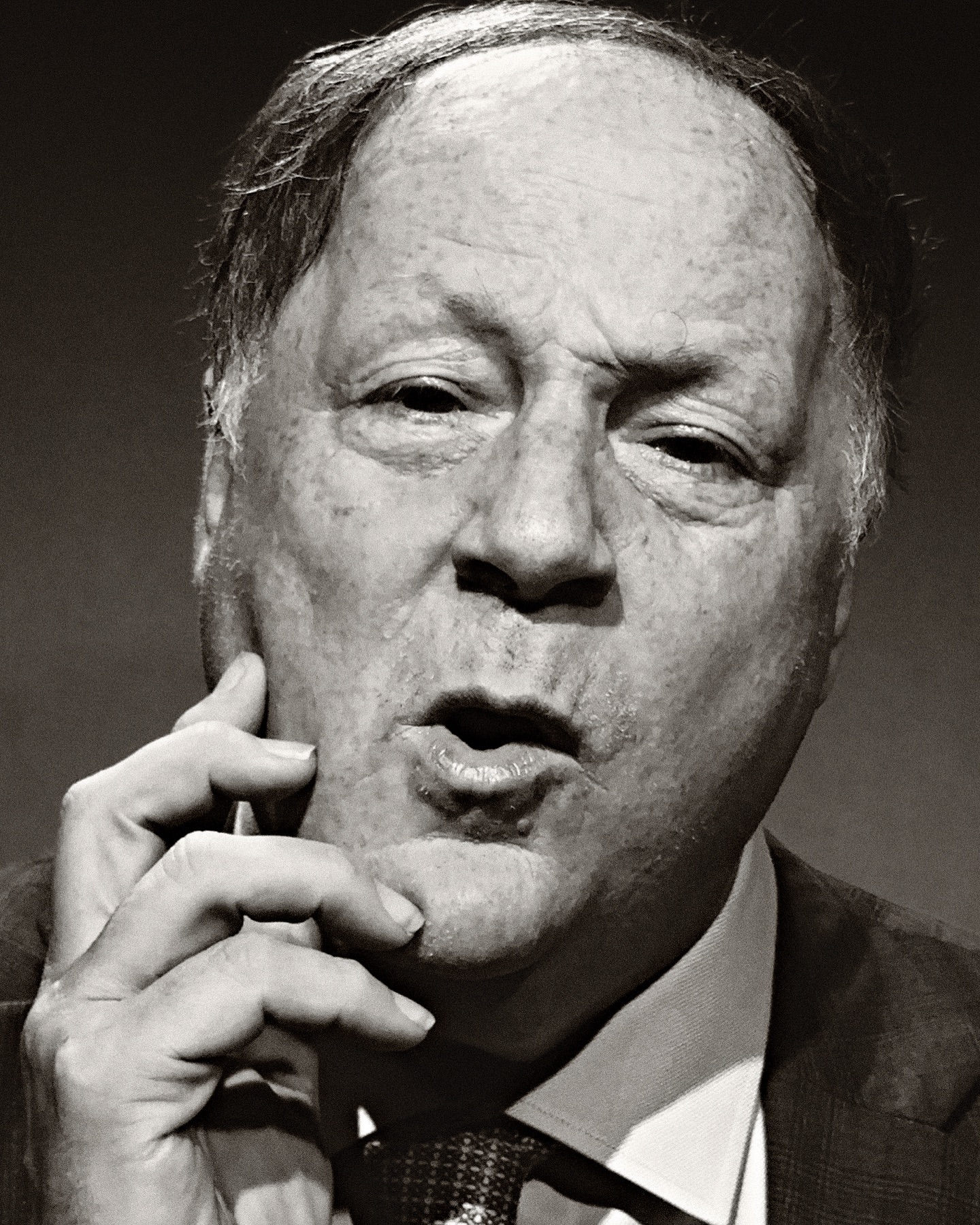 Portrait of an adult man with light skin and dark hair in a suit and tie, holding his hand to his face in a pensive pose.