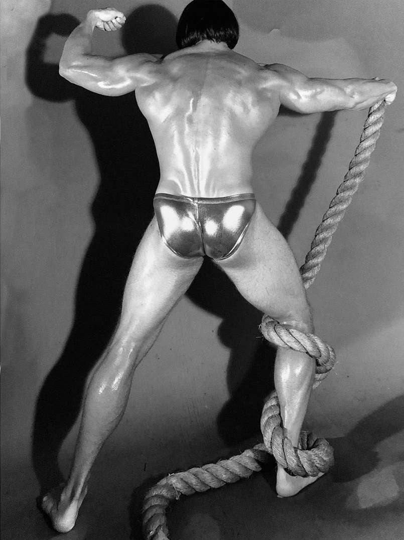 Rear view of a muscular man in a metallic bathing suit posing with a thick rope, flexing his bicep in a monochrome studio shot.
