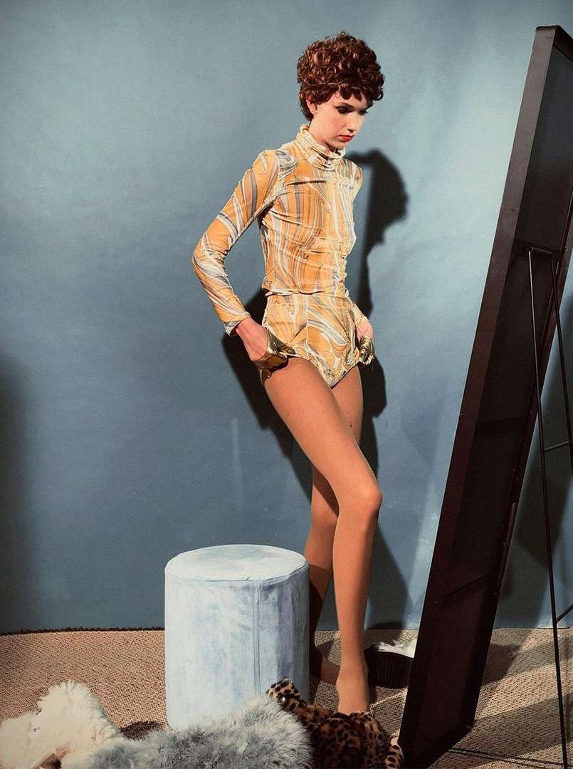 A fashion-style photo showcases an adult female model in a gold-patterned two-piece outfit, standing next to a mirror and a blue stool.