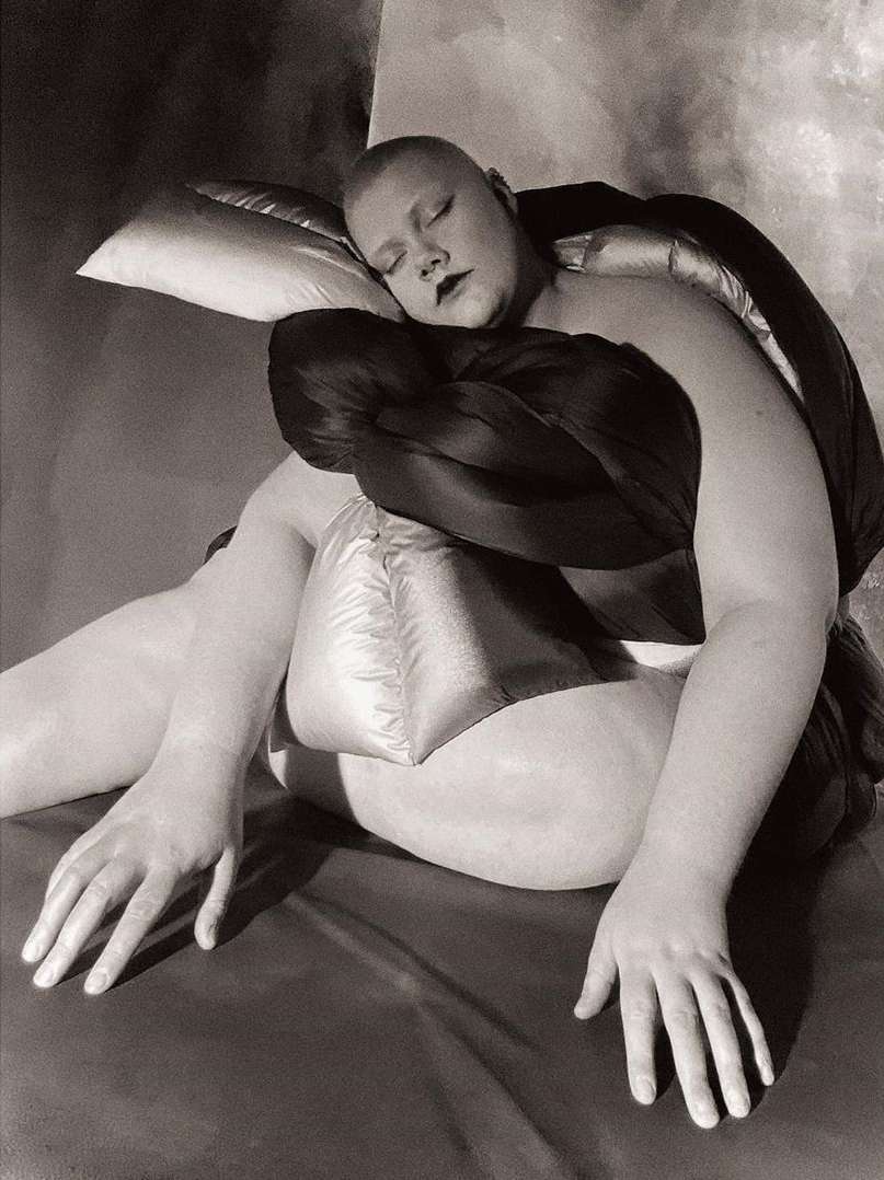 Black and white portrait of a bald person draped in fabric lying on a smooth surface with a satin pillow, sleeping peacefully.