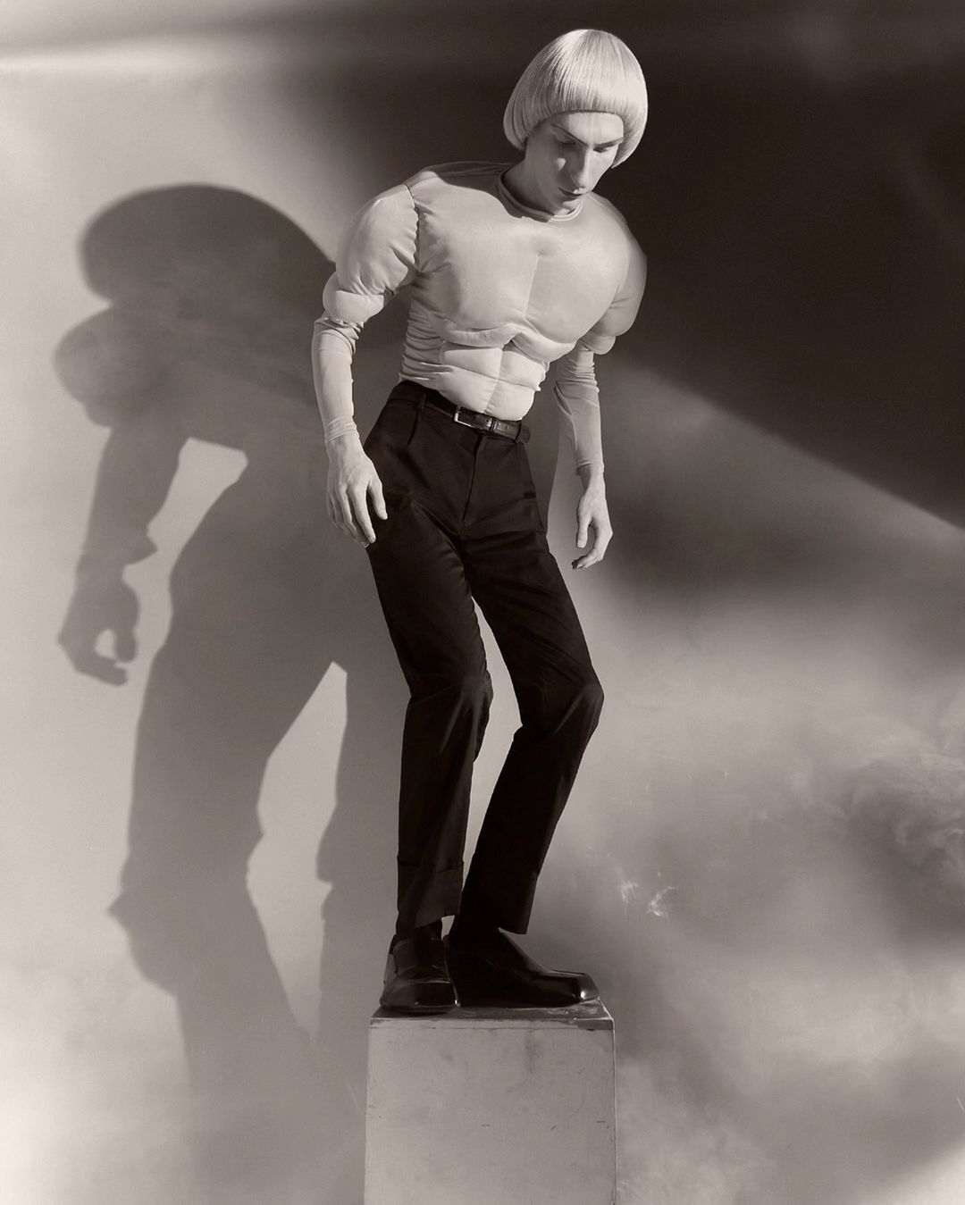 Monochrome studio shot of a slender adult in black pants, boots, and a muscular padded top, standing on a pedestal, with a cast shadow.