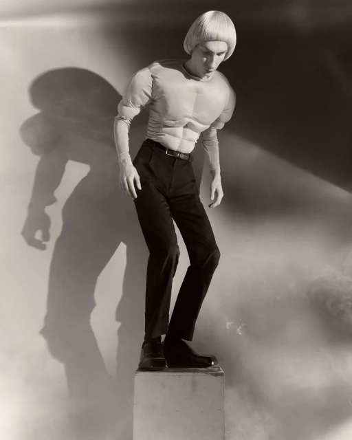 Monochrome studio shot of a slender adult in black pants, boots, and a muscular padded top, standing on a pedestal, with a cast shadow.