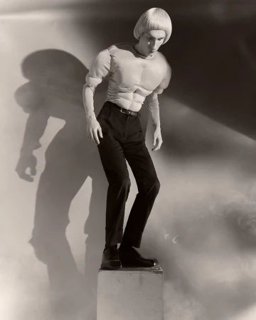 Monochrome studio shot of a slender adult in black pants, boots, and a muscular padded top, standing on a pedestal, with a cast shadow.