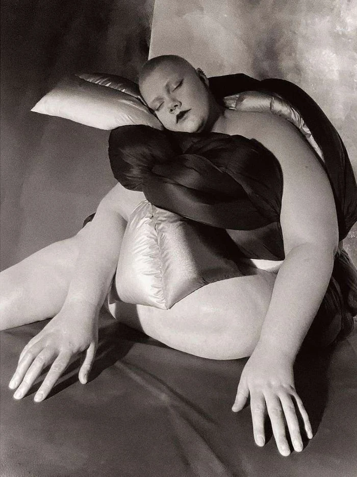 Monochrome portrait by Paul Kooiker of a bald person resting among pillows wrapped in dark and metallic fabric.