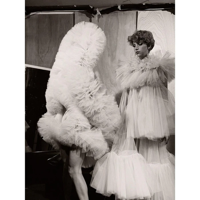 Model in voluminous white tulle ruffles standing beside a sculptural layered ruffle garment against a wooden paneled wall