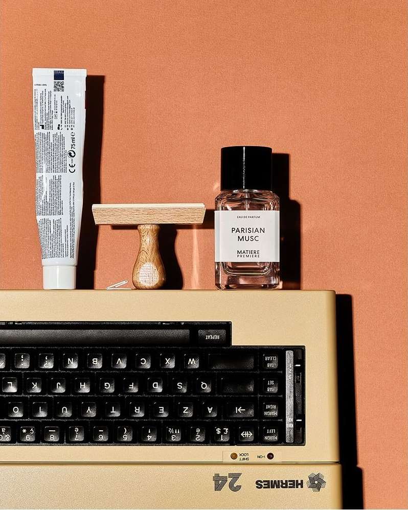 Still life shot of a keyboard, perfume bottle, moisturizer tube, and wooden stamp against an orange backdrop, in a studio setting.