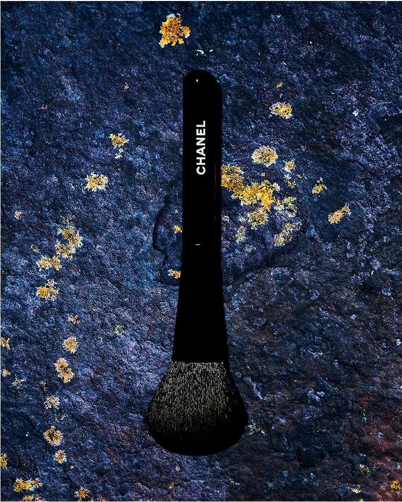 Black Chanel makeup brush on dark blue textured rock surface. The makeup accessory is lit from above in a close-up shot.