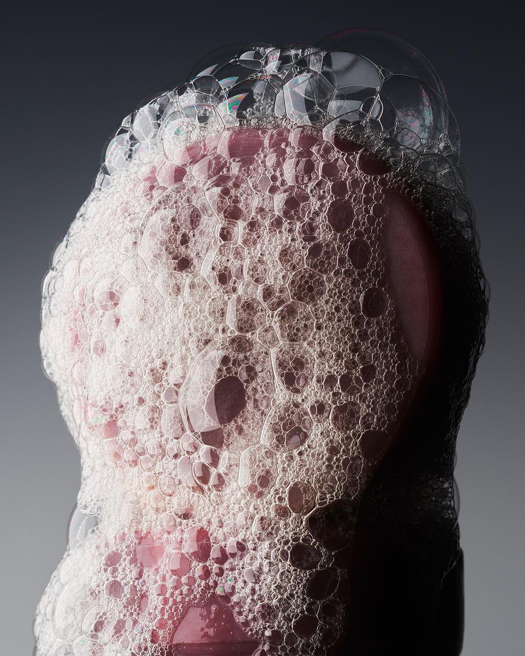 Close-up of a pink object covered in white and clear soap bubbles against a grey background, focusing on texture and light.