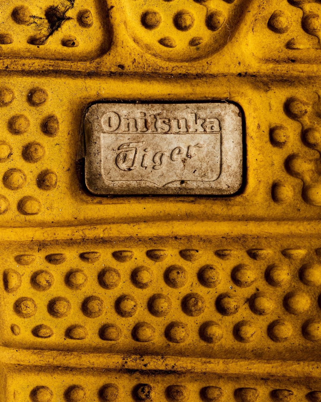 Close-up on an Onitsuka Tiger brand logo plate affixed to yellow patterned material; textured surface, mottled appearance, studio lighting.