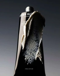 Close-up product shot of a dark skincare tube smeared with a thick beige exfoliating cream against a gray gradient background.